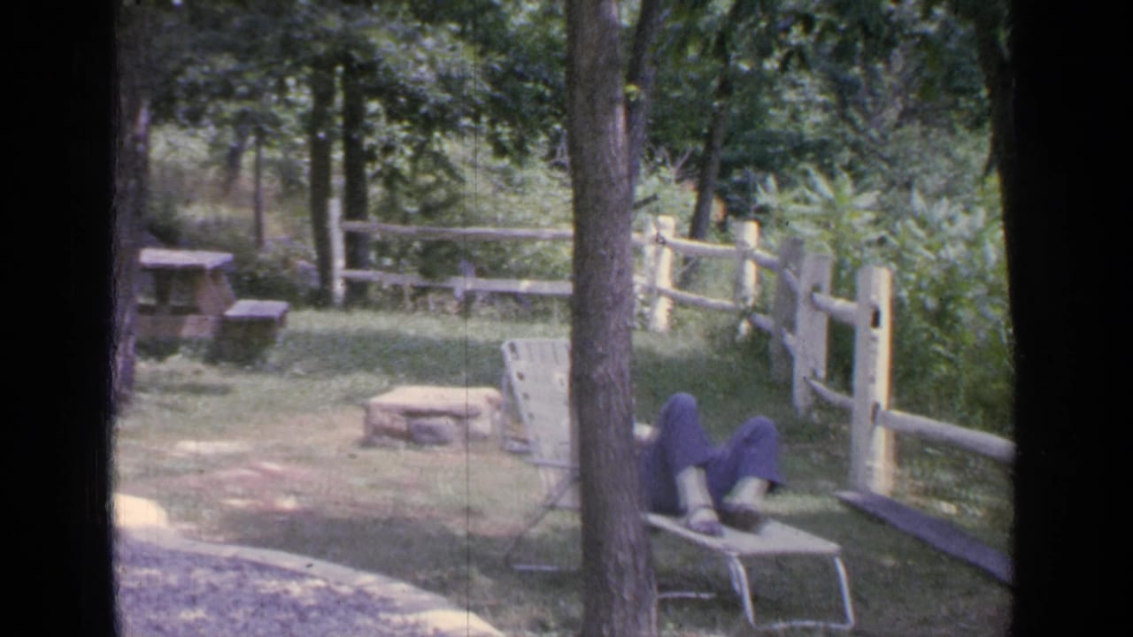 Woman seated on a metal lounge chair in a wooded yard near a split-rail fence.