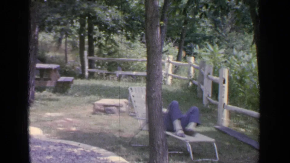 1964 Greenwood Lake home-movie frame showing a woman resting on a metal lounge chair beside trees, grass, and a rustic fence.