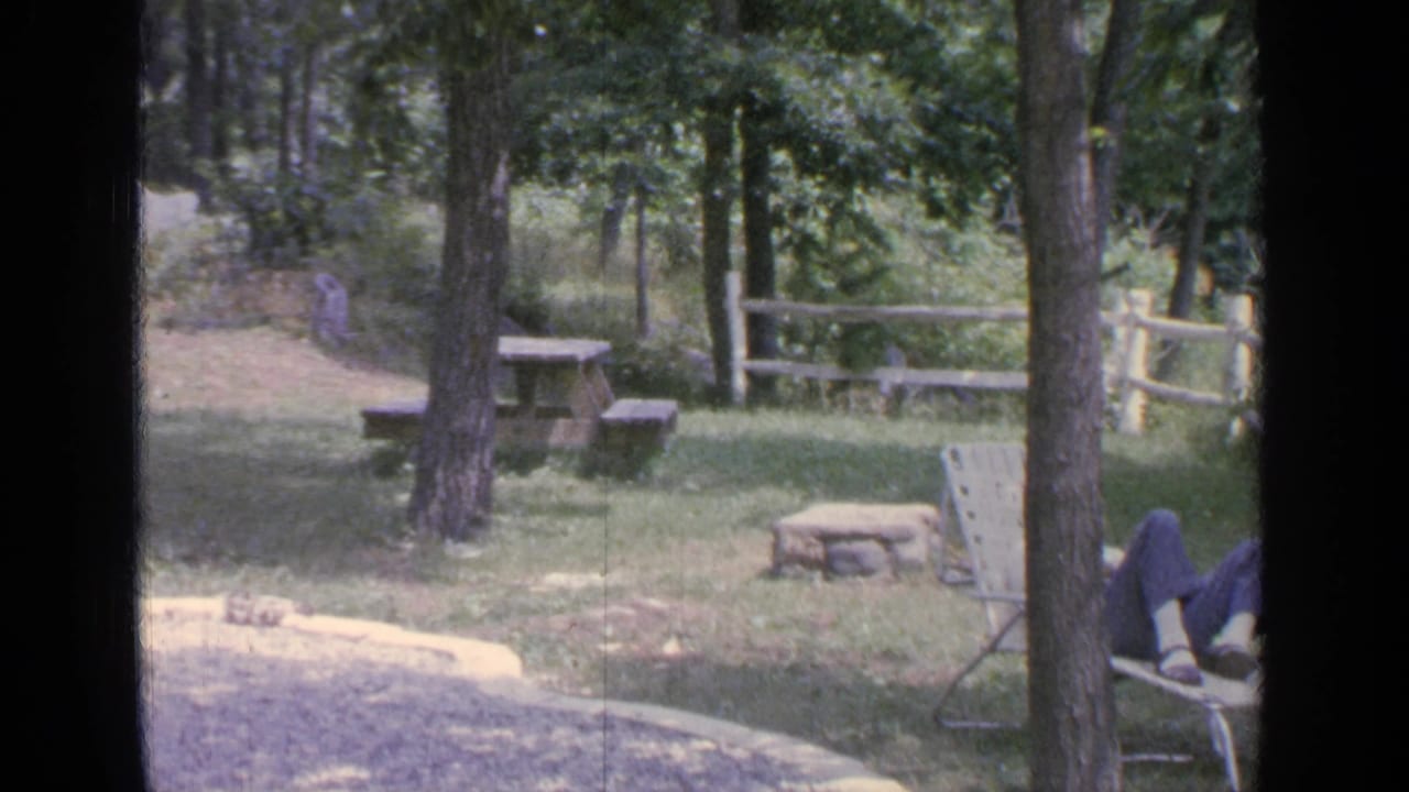 1964 Greenwood Lake backyard view with picnic table, split-rail fence, fire pit, trees, and the seated woman partly cropped at right.