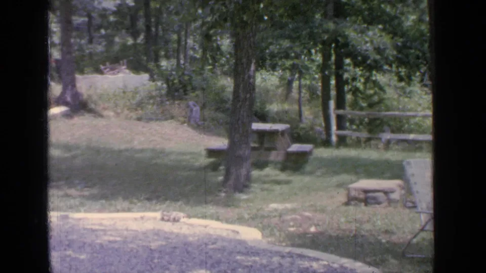 1964 Greenwood Lake home-movie frame of a shaded yard with a central tree, picnic table, rustic fence, fire pit, and partial lounge chair.