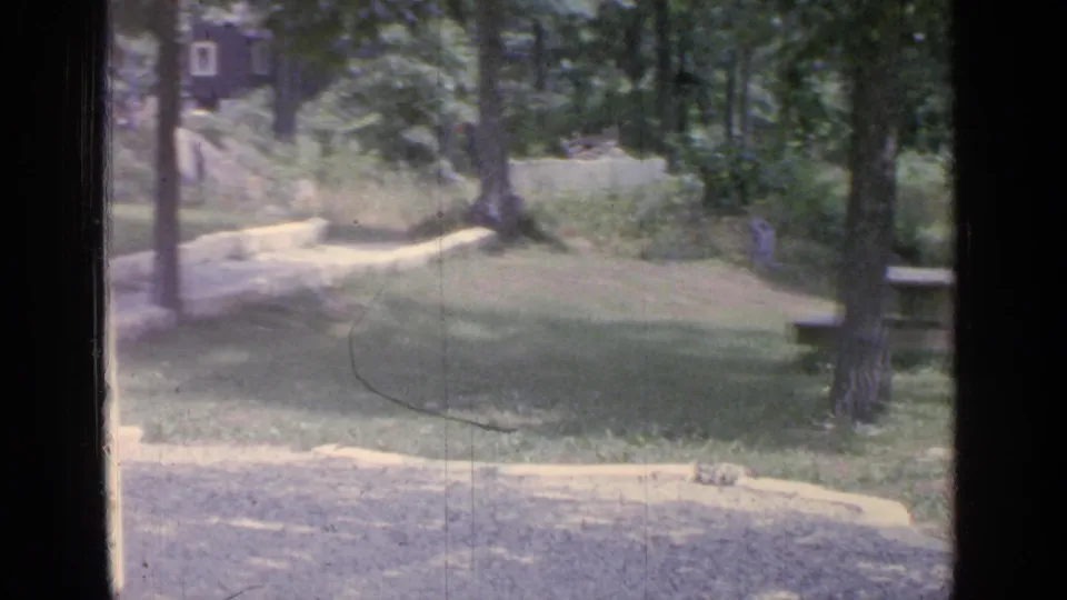 1964 8mm view of a shaded Greenwood Lake park area with trees, grass, a light path, and a small distant building.