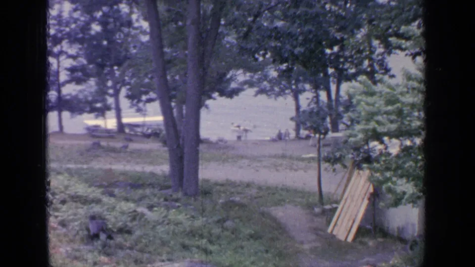 1964 lakeside park view at Greenwood Lake with shaded ground, trees, picnic tables, distant people, and shoreline water beyond.