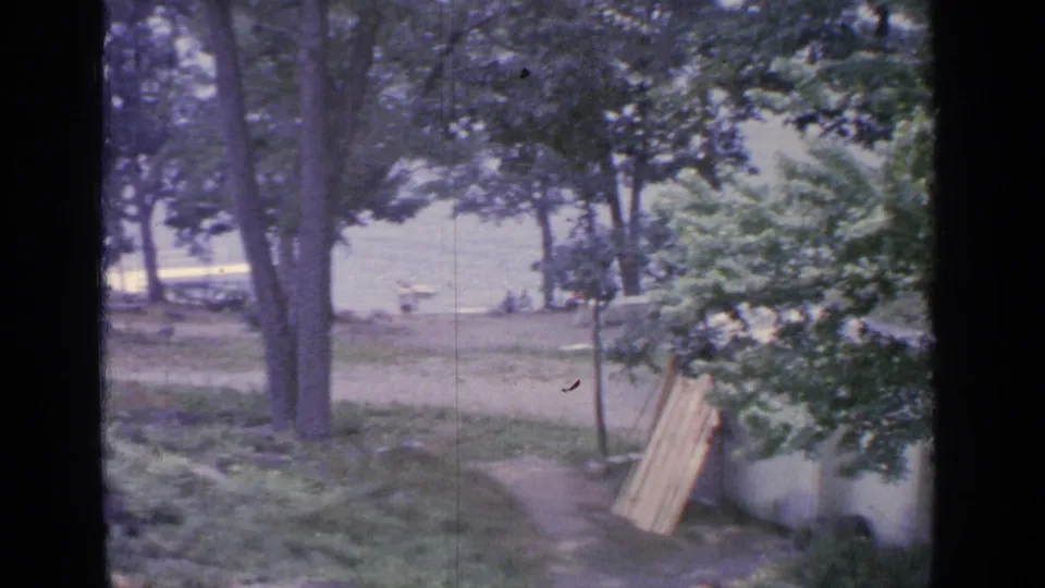 Static 8mm frame of Greenwood Lake parkland with trees, a path, distant waterfront visitors, and boards leaning near a fence.
