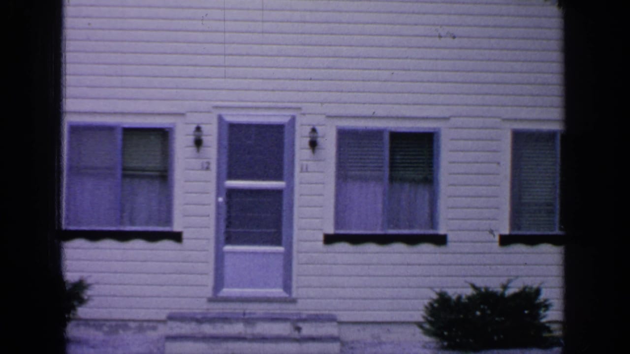 White clapboard house exterior with windows, blinds, a door, and bushes near the base.