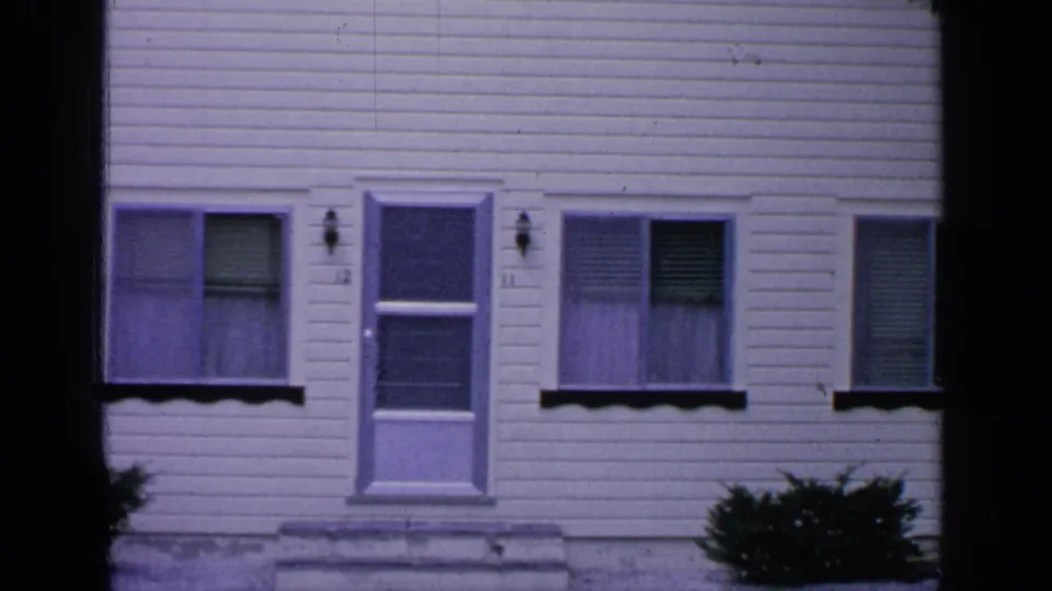 1964 Greenwood Lake home-movie frame showing a white clapboard house facade with windows, blinds, a door, and shrubs.