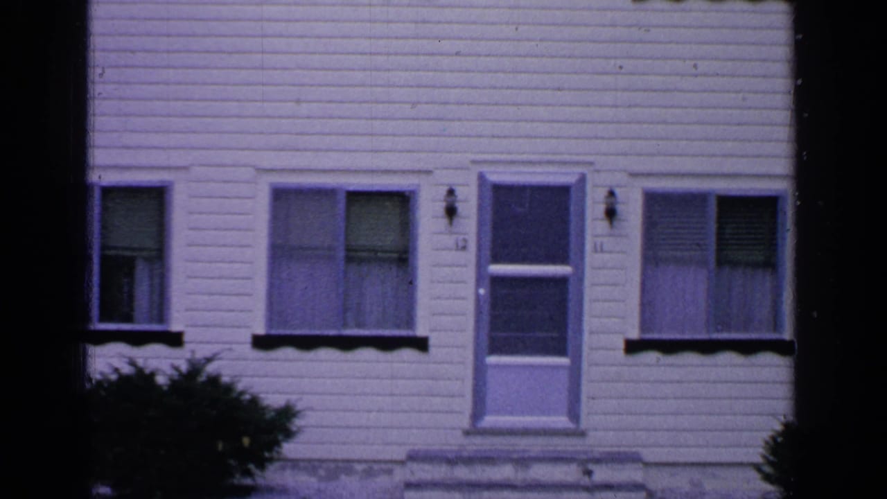 Closer view of the white house facade with a door, windows, blinds, and bushes.
