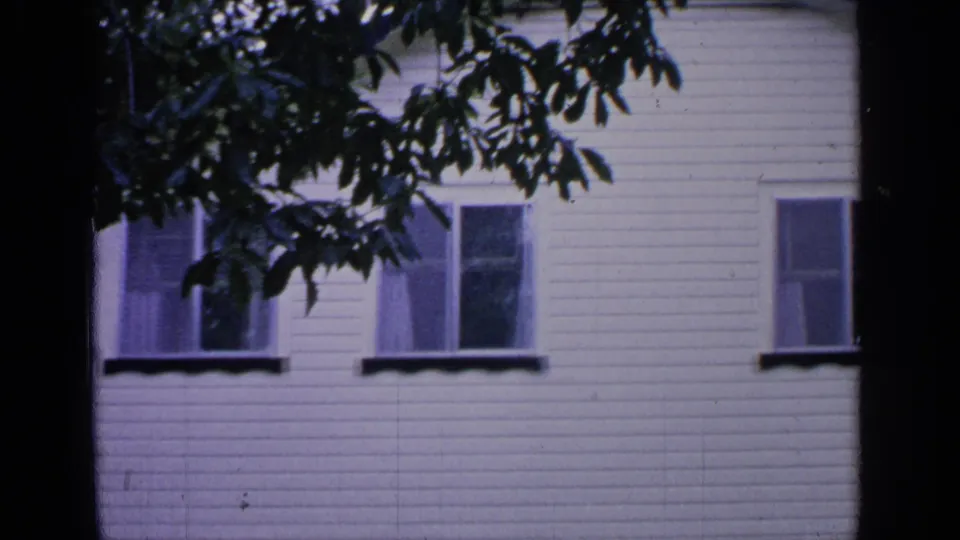 1964 8mm view in Greenwood Lake, New York, showing the upper portion of a white clapboard residential facade behind tree branches.