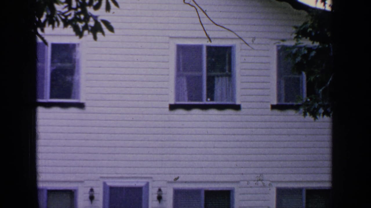 Greenwood Lake home-movie frame with a white clapboard residential facade, multiple windows, and a dark tree trunk crossing the center.