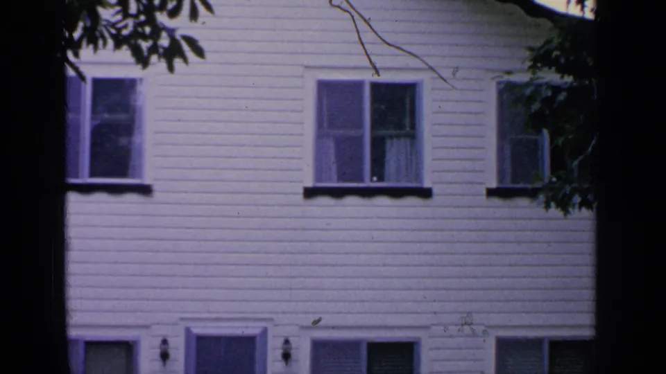 Greenwood Lake home-movie frame with a white clapboard residential facade, multiple windows, and a dark tree trunk crossing the center.