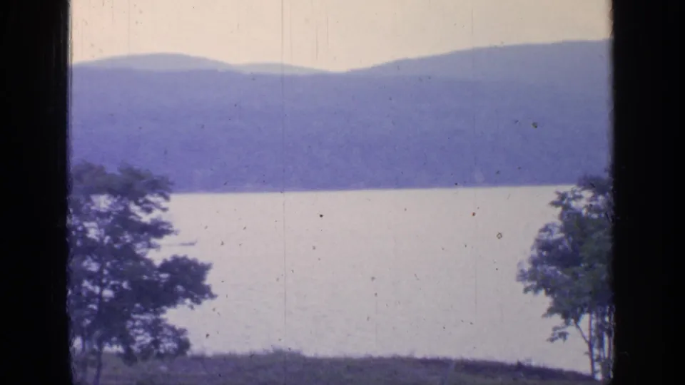1964 Greenwood Lake home-movie frame showing bright water, blue-gray hills, and low shoreline trees at the frame edges.