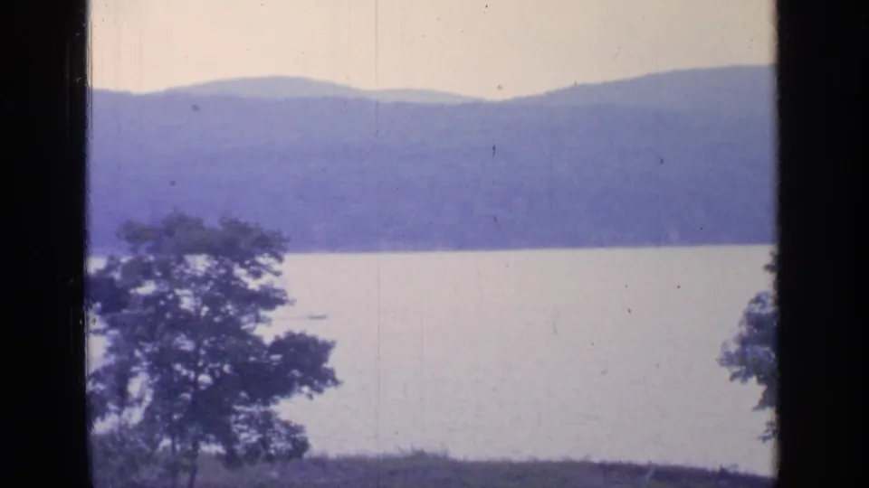 1964 Greenwood Lake view with foreground trees, calm water, distant wooded mountains, and dark vertical frame edges.