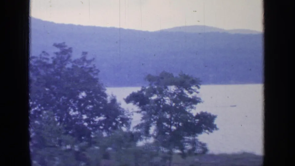 1964 Greenwood Lake view with trees in the foreground, a small distant boat on the water, and hazy mountains across the lake.