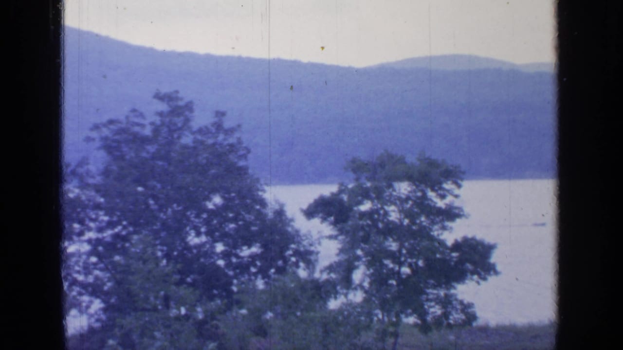 1964 Greenwood Lake view with foreground trees, pale water, distant wooded hills, and dark window bars in an 8mm frame.