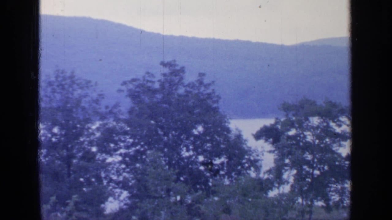 Window-framed 8mm view of Greenwood Lake, with leafy trees in front of pale water and blue-toned mountain ridges.