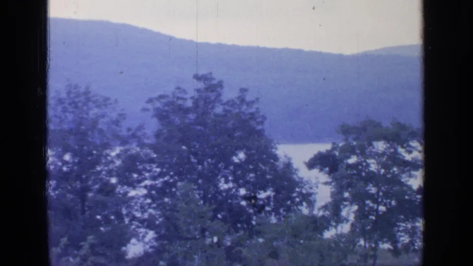 Window-framed 8mm view of Greenwood Lake, with leafy trees in front of pale water and blue-toned mountain ridges.