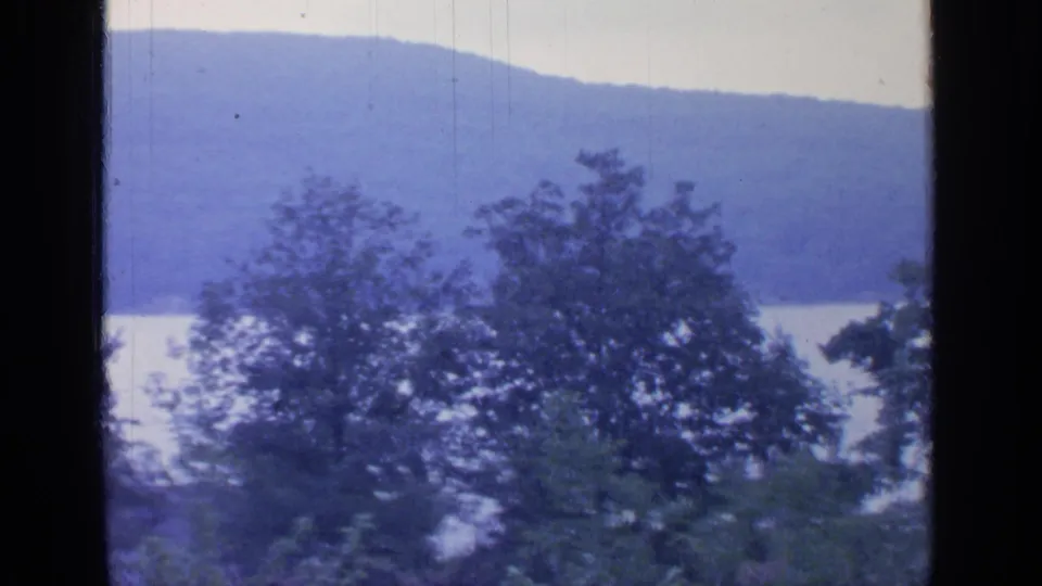 1964 home-movie frame showing Greenwood Lake through a window, with trees, calm water, and a low blue ridge beyond.