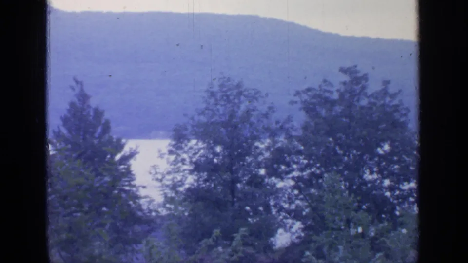 Blue-toned 8mm frame of Greenwood Lake scenery, with trees in the foreground and hazy wooded hills across the water.