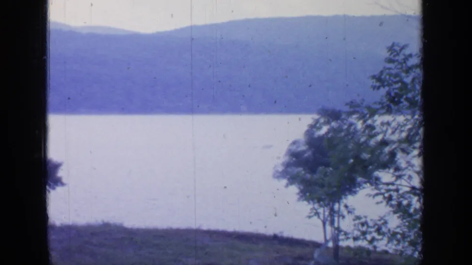 1964 Greenwood Lake home-movie frame showing calm water, wooded hills, shoreline trees, pale sky, and dark film or window borders.
