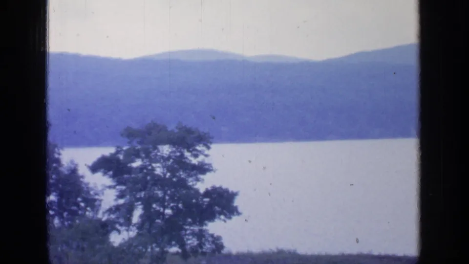 1964 Greenwood Lake scenery with blue hills across the water, a broad tree silhouette in front, and visible 8mm grain and scratches.