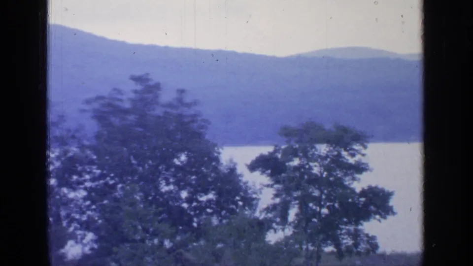 A later frame from the Greenwood Lake clip shows trees along the water with blue mountain slopes beyond and a dark vertical border at left.