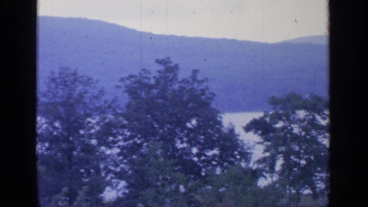 1964 8mm view likely at Greenwood Lake, New York, showing trees, pale water, and blue mountains beyond a dark window frame.
