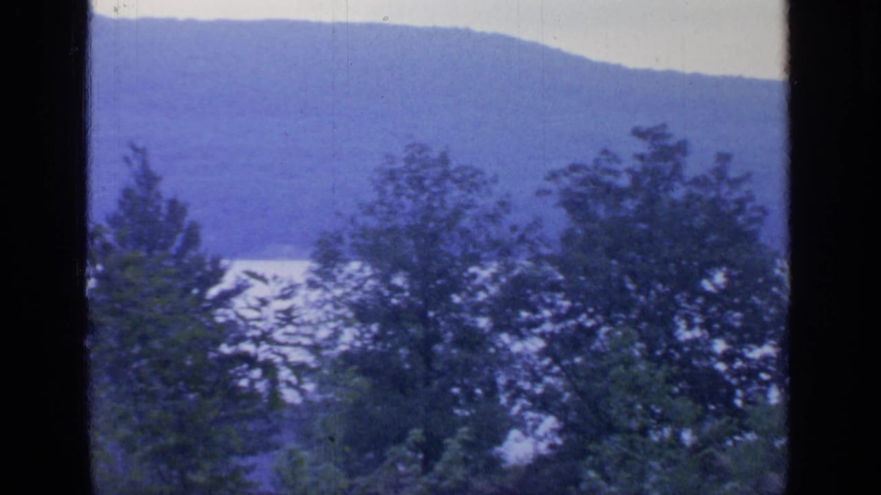 Likely Greenwood Lake landscape from 1964, with leafy trees before pale water and a soft blue mountain ridge.