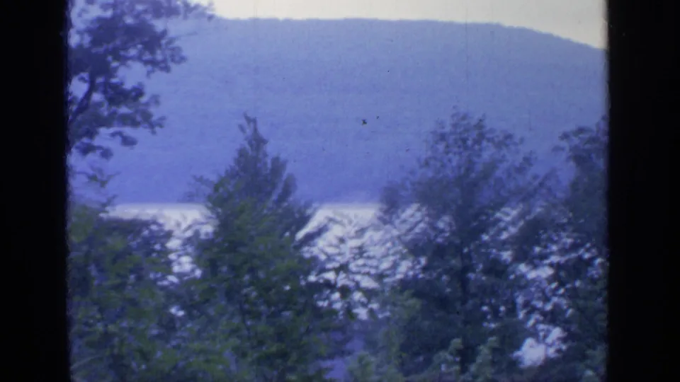 1964 8mm landscape view likely at Greenwood Lake, with dense foreground trees, bright water, and hazy mountains in the distance.