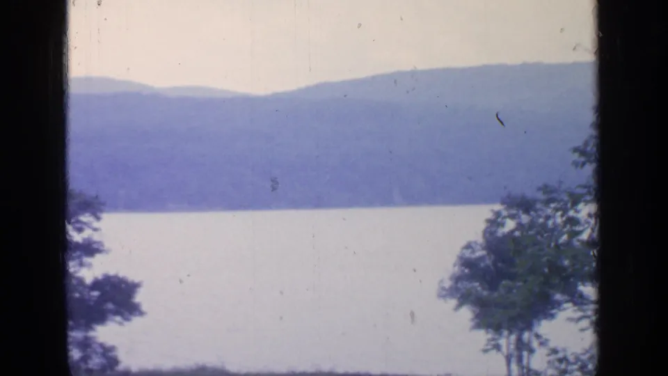 1964 8mm view across Greenwood Lake toward distant wooded hills, with trees along the lower and side edges.