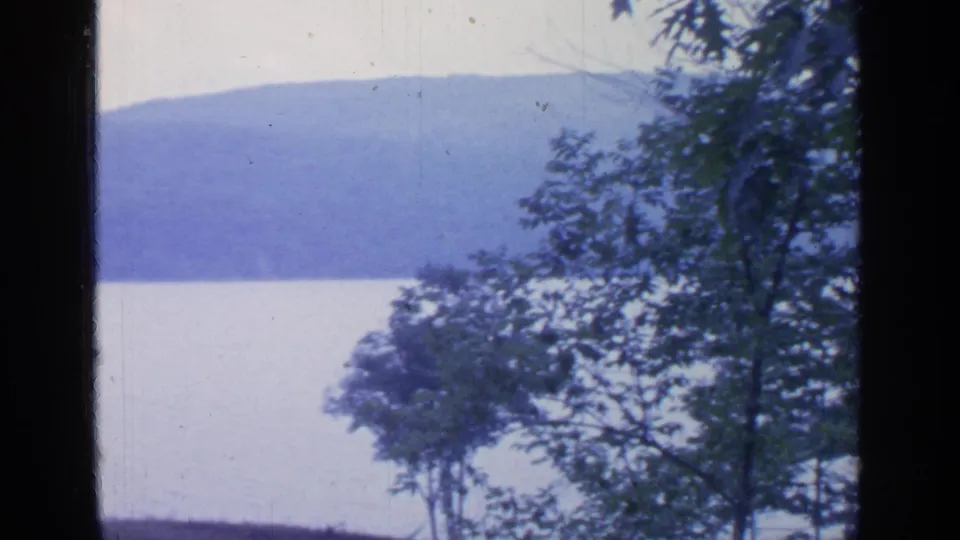 Greenwood Lake seen through leafy foreground trees, with pale water and hazy blue hills in the distance.