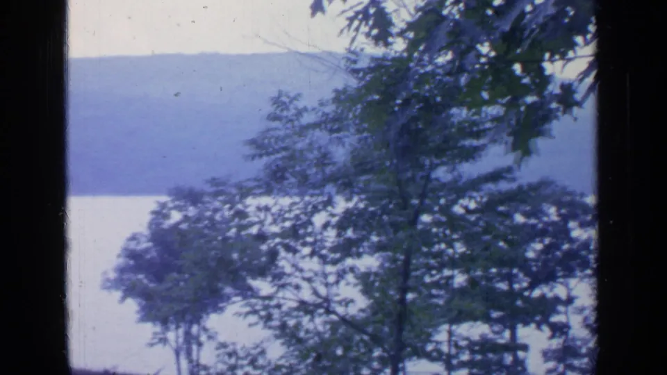 1964 Greenwood Lake view through dark framing, with trees in the foreground and a calm lake and wooded hills in the distance.