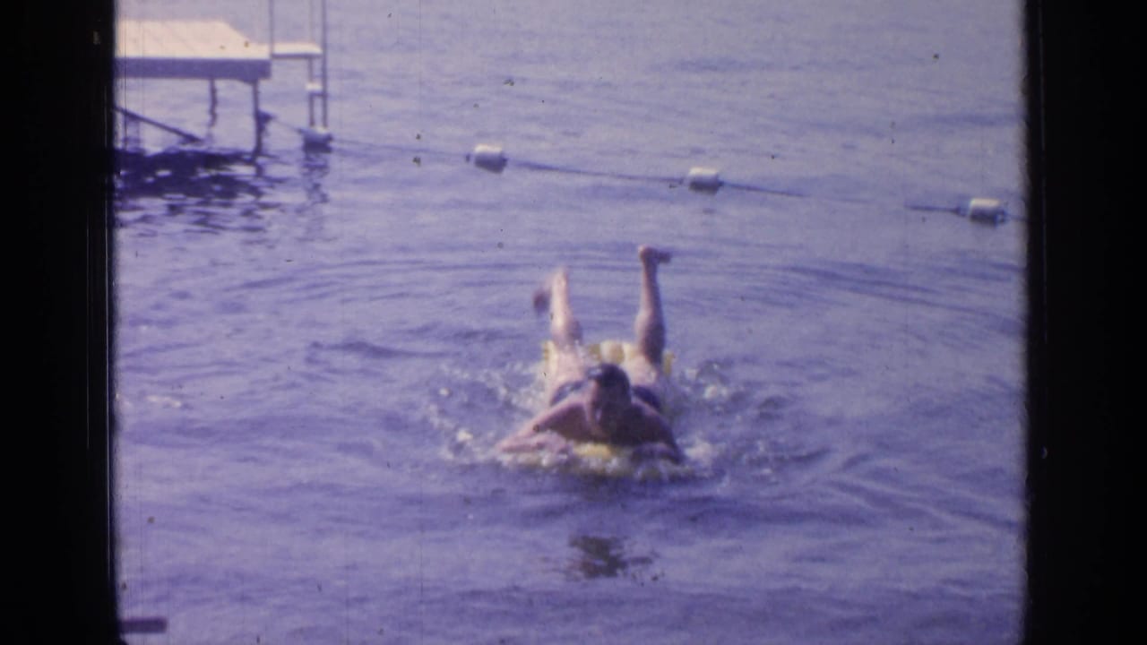 Man swimming toward the camera on a yellow inflatable ring in lake water near buoys.