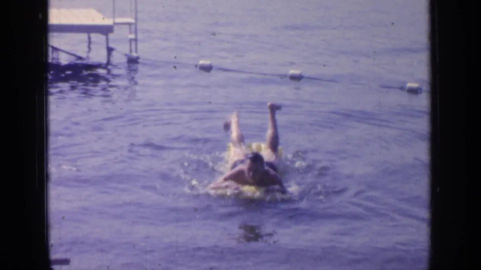 1964 Greenwood Lake home-movie frame showing a man kicking through the water on a yellow inflatable ring near a buoy line.