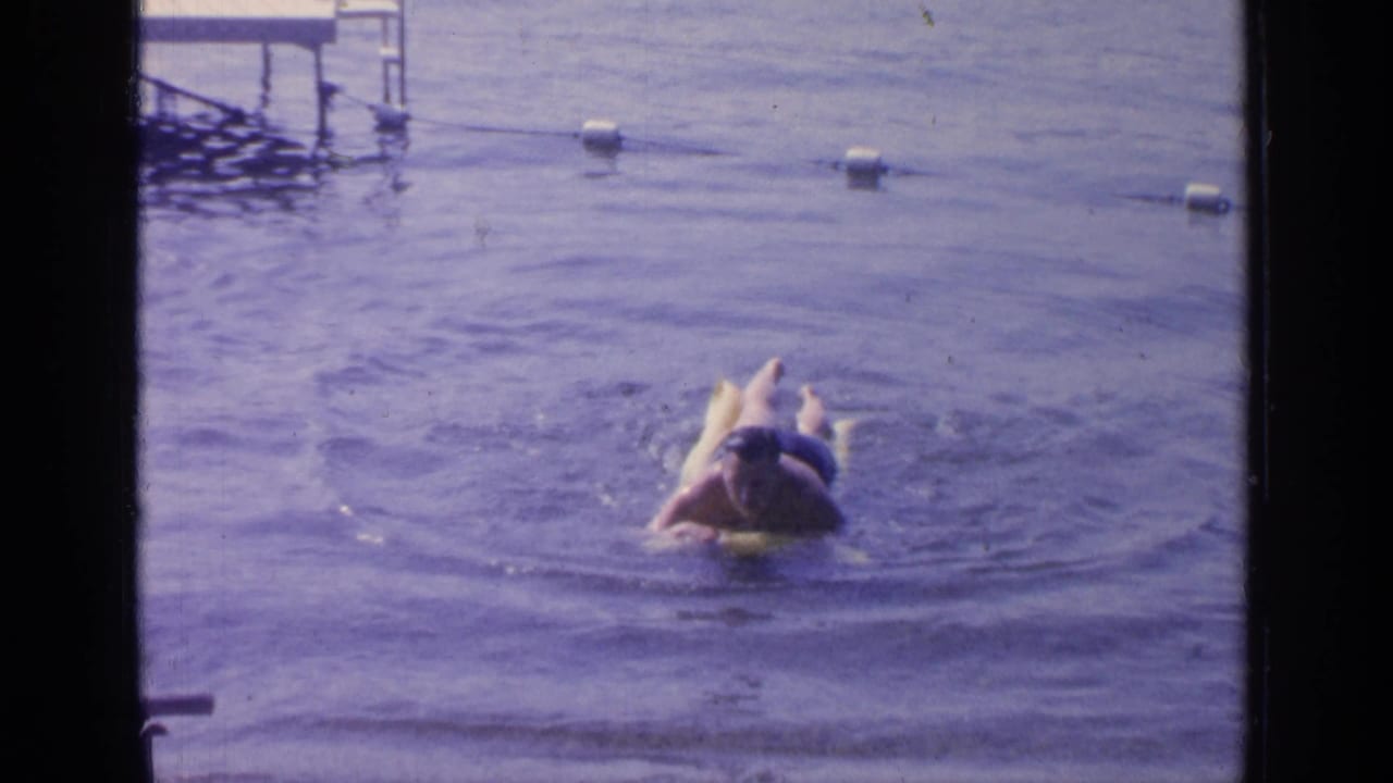 1964 Greenwood Lake frame with a man lying on a yellow inflatable ring, legs visible as ripples spread across the water.