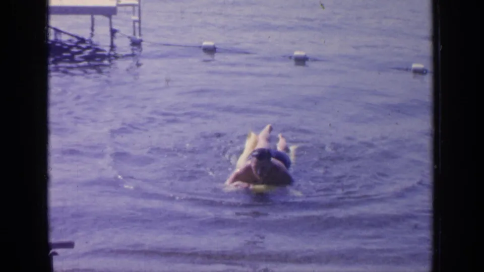 1964 Greenwood Lake frame with a man lying on a yellow inflatable ring, legs visible as ripples spread across the water.