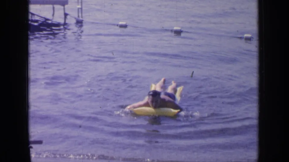 1964 Greenwood Lake home-movie frame showing a man on a yellow inflatable ring moving through the water below a line of white buoys.