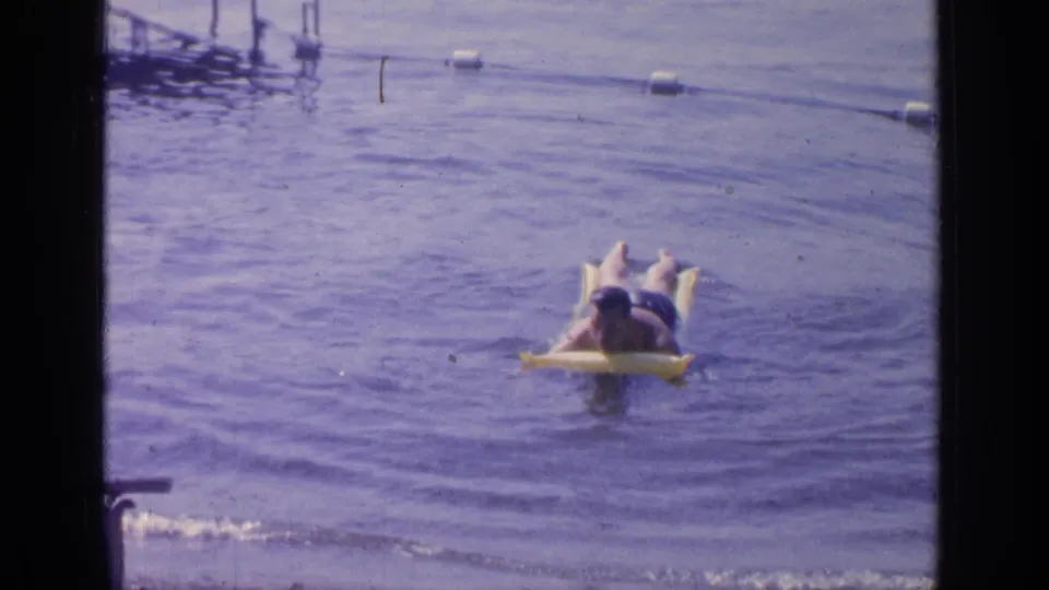 1964 Greenwood Lake frame of a man floating on a yellow inflatable ring in calm water, with buoys and dock details behind him.