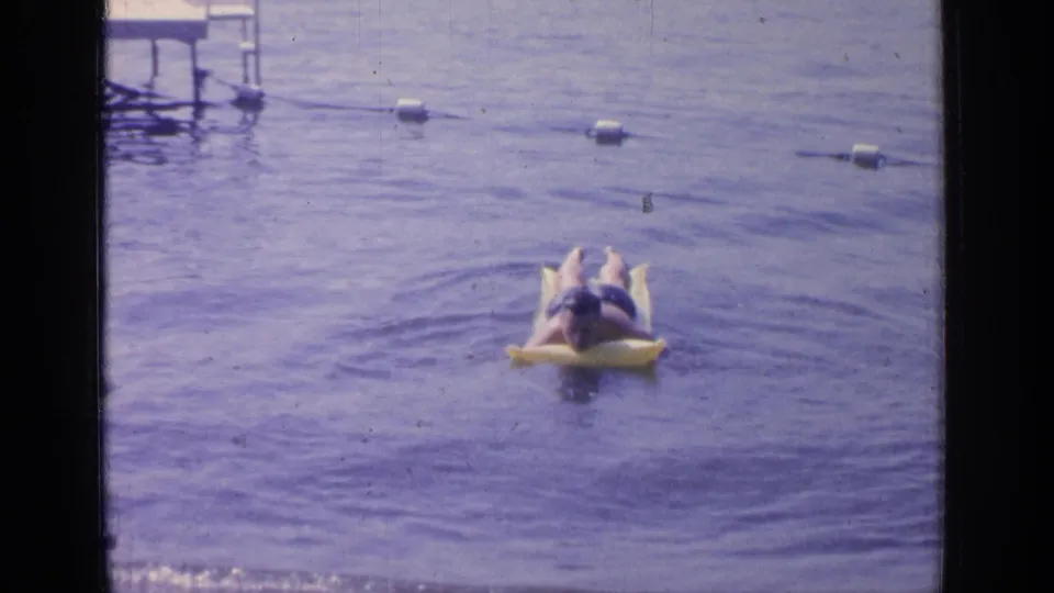 1964 Greenwood Lake home-movie frame showing a man reclining on a yellow float inside a buoy-marked swimming area.