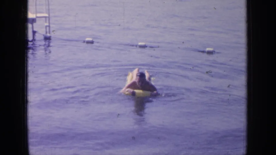 A man on a yellow inflatable float faces toward the camera in the roped Greenwood Lake swimming area.