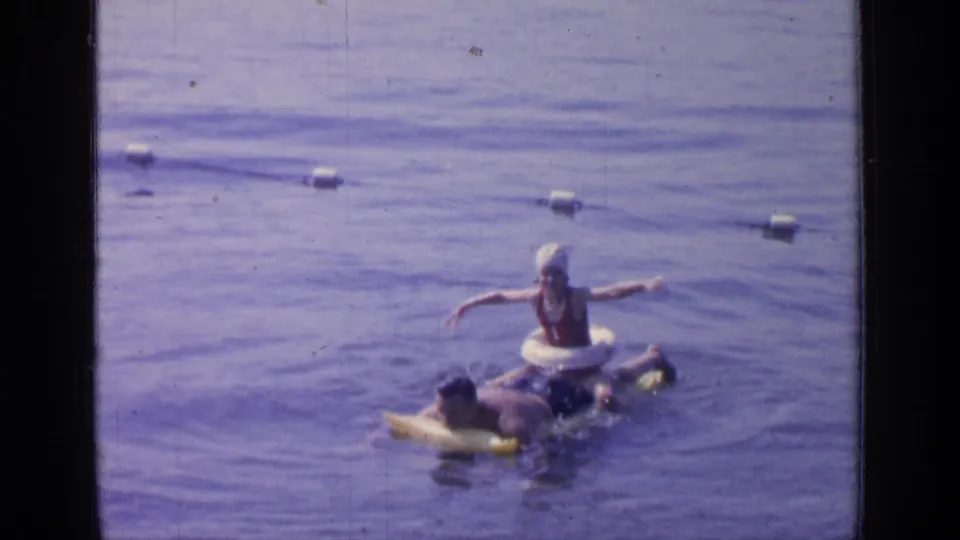 A woman in a white swim cap joins the man on inflatables in the Greenwood Lake swimming area, filmed in 1964.