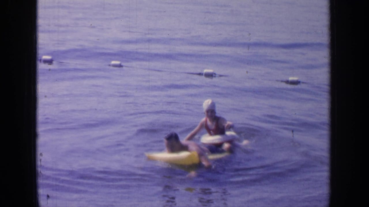 At Greenwood Lake in 1964, two adults play around a yellow inflatable raft in blue water, with a buoy line visible behind them.