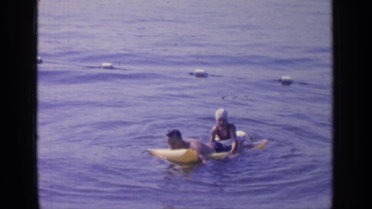 A 1964 Greenwood Lake home-movie frame shows a swimmer on a yellow inflatable raft interacting with a woman in a white swim cap.