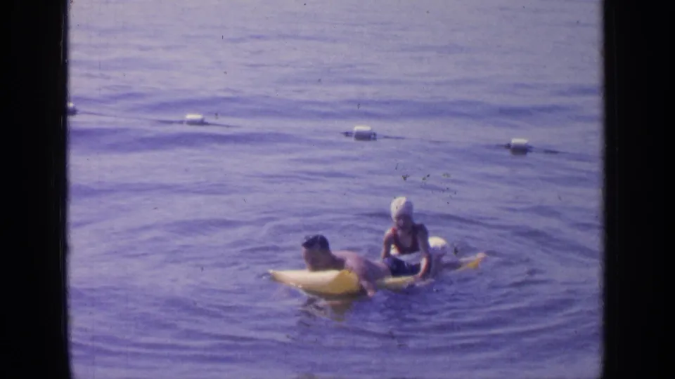 A 1964 Greenwood Lake home-movie frame shows a swimmer on a yellow inflatable raft interacting with a woman in a white swim cap.