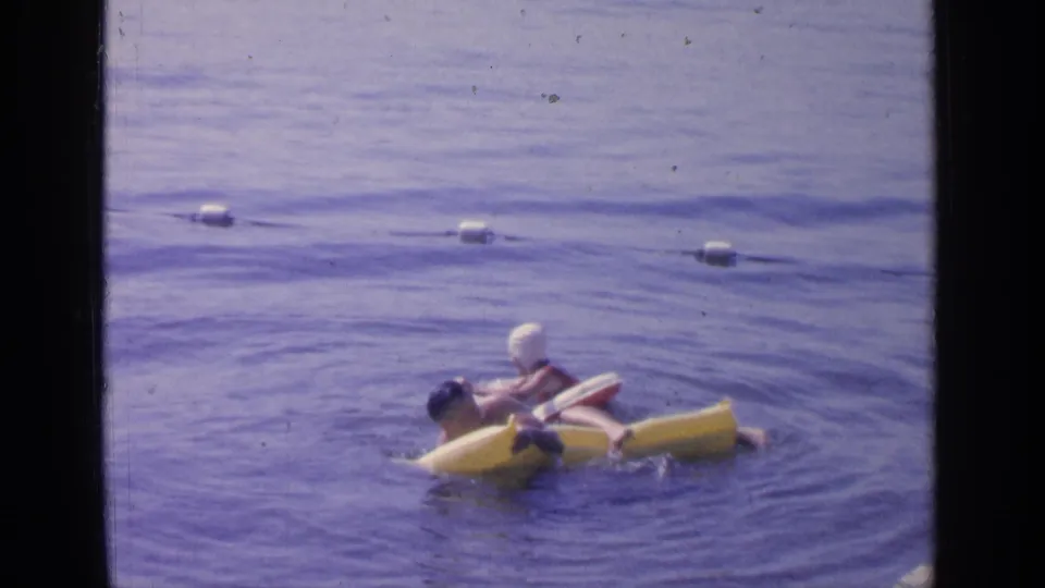 Two adults at Greenwood Lake in 1964 appear to jostle and balance around a yellow inflatable raft in a casual swimming scene.