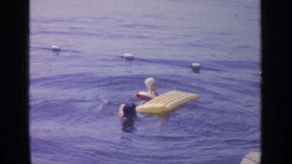At Greenwood Lake in 1964, two swimmers are in calm water near a yellow inflatable raft and white buoys.