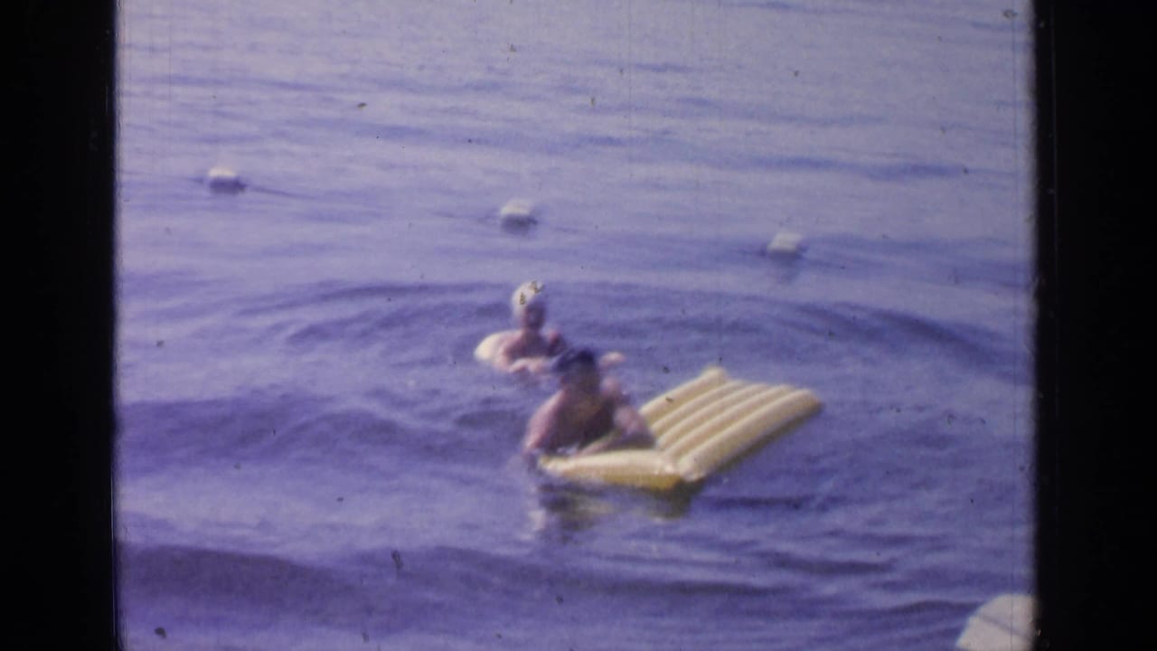 Two people swim at Greenwood Lake in 1964, with one holding a yellow inflatable raft near a buoy line.