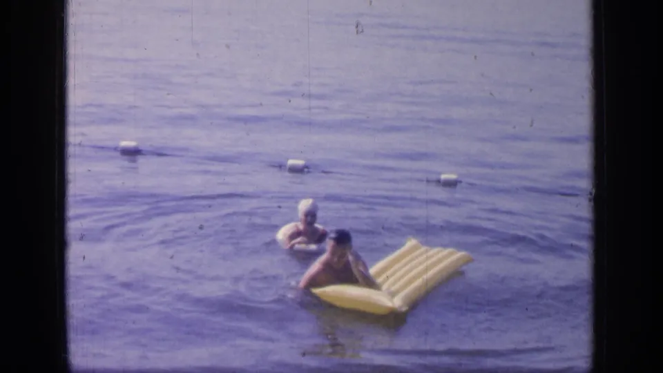 A swimmer leans on a yellow raft at Greenwood Lake as another person in a swim cap stays close by.