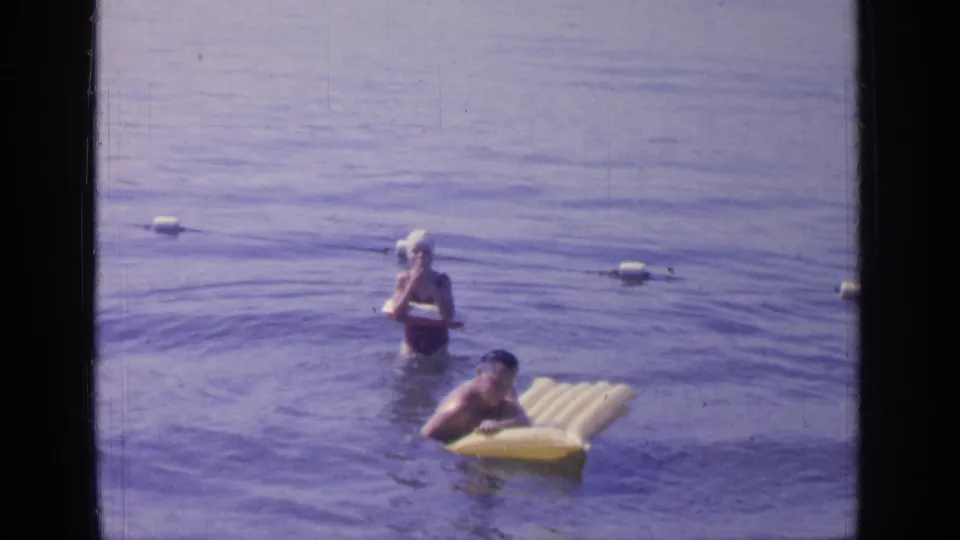 In the 1964 Greenwood Lake footage, one swimmer rests on a yellow raft while another stands in shallow water behind.