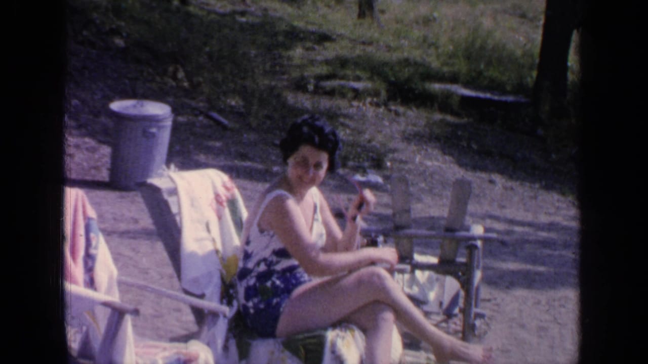 Woman in a floral swimsuit sits on a beach chair beside towels and a metal trash can.