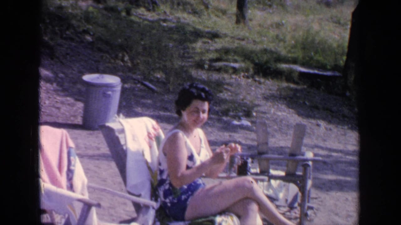 At Greenwood Lake in 1964, the woman looks toward the camera while seated in a beach chair, with towels and a metal trash can nearby.