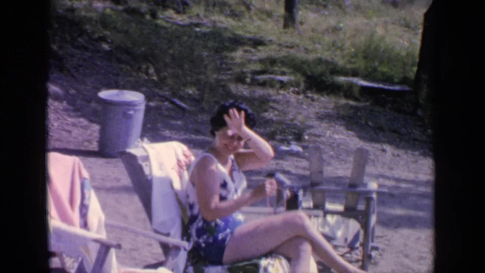 At Greenwood Lake in 1964, the seated woman raises her hand toward her face or the camera while resting in a beach chair.